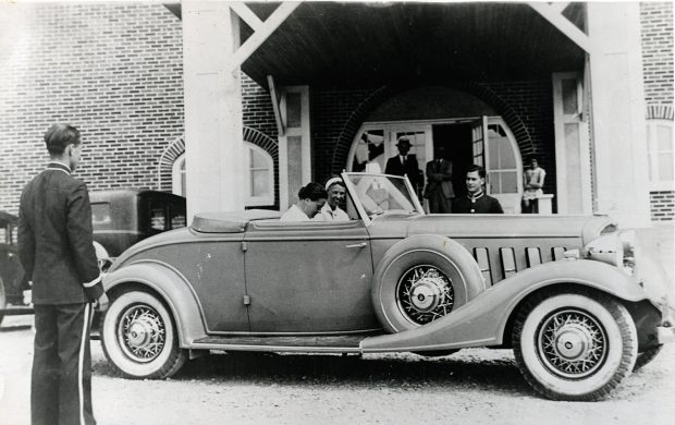 Black and white photograph. In a convertible Buick, two women, Eleanor Roosevelt and her friend Lorena Hickok, are about to depart. The car is parked at the front of the La Belle Plage hotel in Matane. A doorman and bellboy from the hotel are waiting for the departure of their distinguished guests.