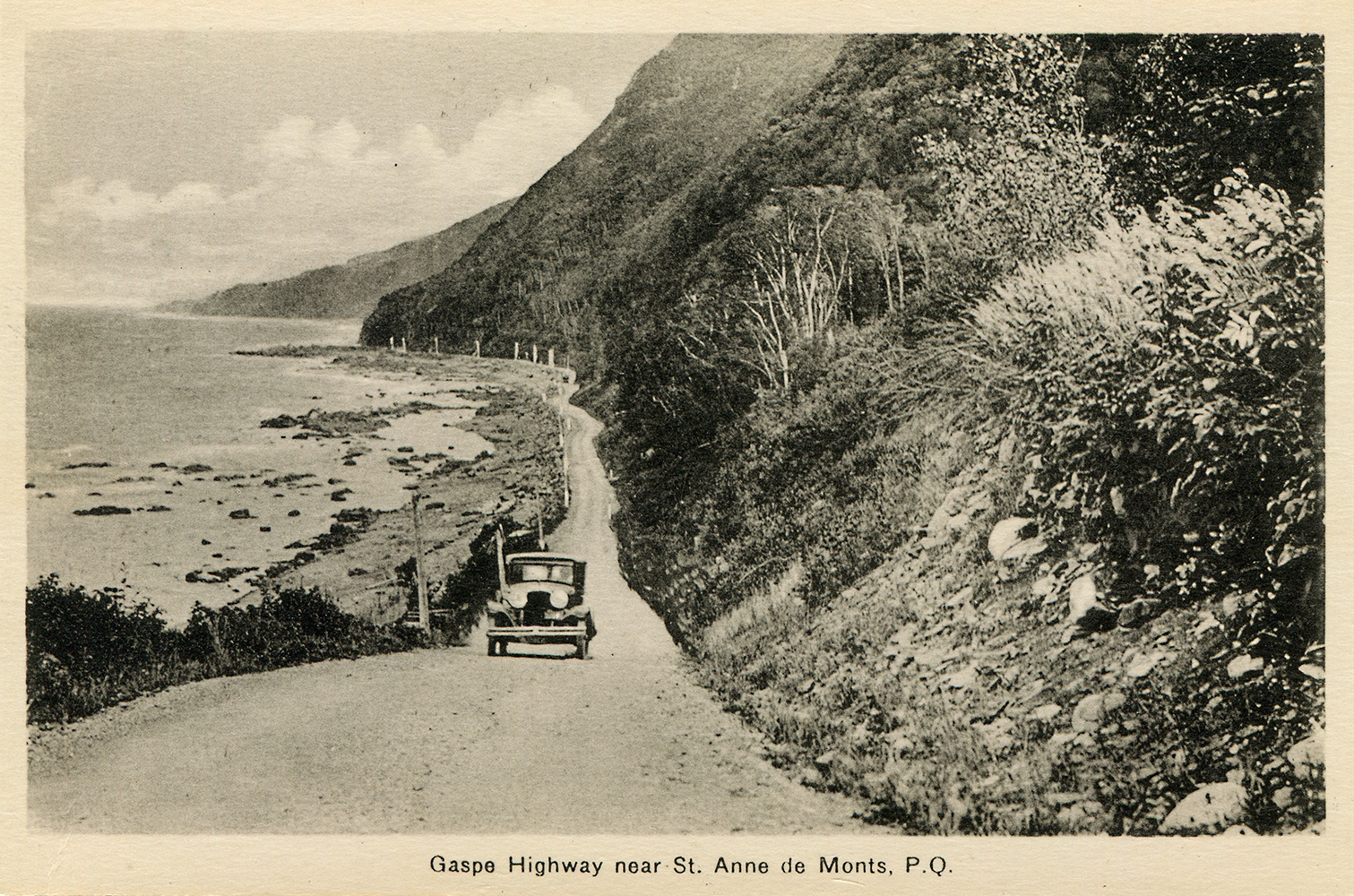 Postcard showing a car rolling on the gravel surface of the Perron Boulevard around 1930. The Perron Boulevard runs along the foot of a steep mountain and the bank of the St. Lawrence River near Sainte-Anne-des-Monts.