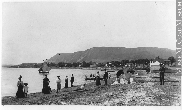 Black and white photograph of a beach. A dozen people pose, men in suits and women in long dresses, some wearing umbrellas. A sailboat and other small boats leave the shore. In the background are some rustic houses at the foot of a mountain.