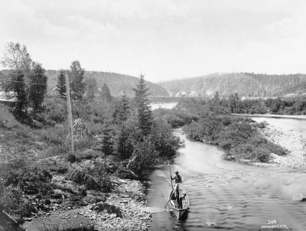 A forest landscape crossed by a river. At the bottom of the photograph, two native men are handling a canoe with poles. On the bank of the river we can see a teepee and, in the distance, some rustic houses at the foot of two hills.