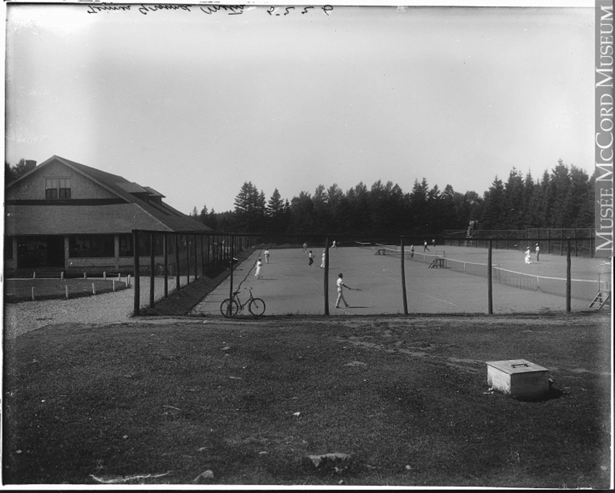 Black and white photograph of a tennis court in Metis Beach. A high fence surrounds the tennis court. Men, wearing white trousers and a white shirt, and women, wearing a long white skirt and a white shirt with long sleeves and hat, are playing tennis. On the left side is the club house of the Cascade Golf and Tennis Club.