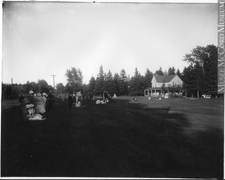 Photographie noir et blanc du départ du parcours du club de golf Cascade. Une vingtaine d’individus vêtue à la mode des années 1914 attendent le départ d’un parcours. Quelques femmes sont assises sur la pelouse. En arrière-plan, un chalet de deux étages à l’architecture victorienne épurée. 