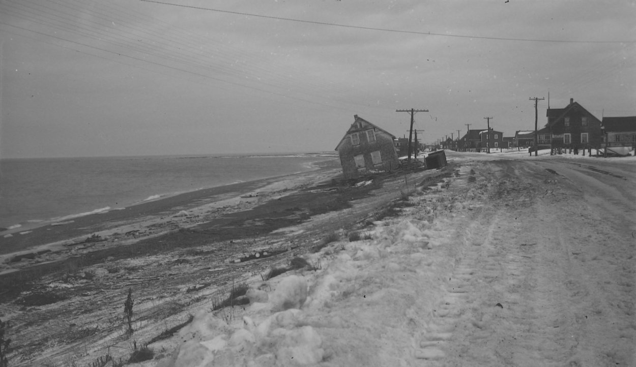 Photograph in black and white of a two-story house at the entrance of a seaside village. The house is on the beach strangely tilted on one side, it appears that it has been moved on the beach side of the road by the waves.