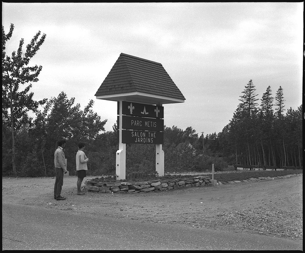 Black and white photograph of the entrance to Parc Metis. A man and a woman observe the sign at the entrance of the site, near Highway 132.