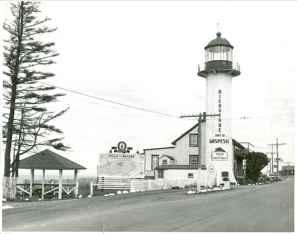 Black and white photograph of the Matane lighthouse, transformed into a tourist information office. Perron Boulevard occupies the foreground of the photograph. The lighthouse and the keeper’s house are located on the right side of the image. On the lighthouse is written: Bienvenue dans la Gaspésie. A poster on the lighthouse mentions: Berceau de la Nouvelle-France. A huge map of Gaspésie is displayed near the lighthouse.