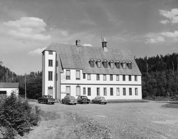 Black and white photograph of the Gîte du Mont-Albert in 1950. The inn is an imposing rectangular building of 3 stories in the heart of a coniferous forest. The pronounced angle of the double slope roof gives it an architectural style that is somewhere between that of a Swiss chalet and a manor house.