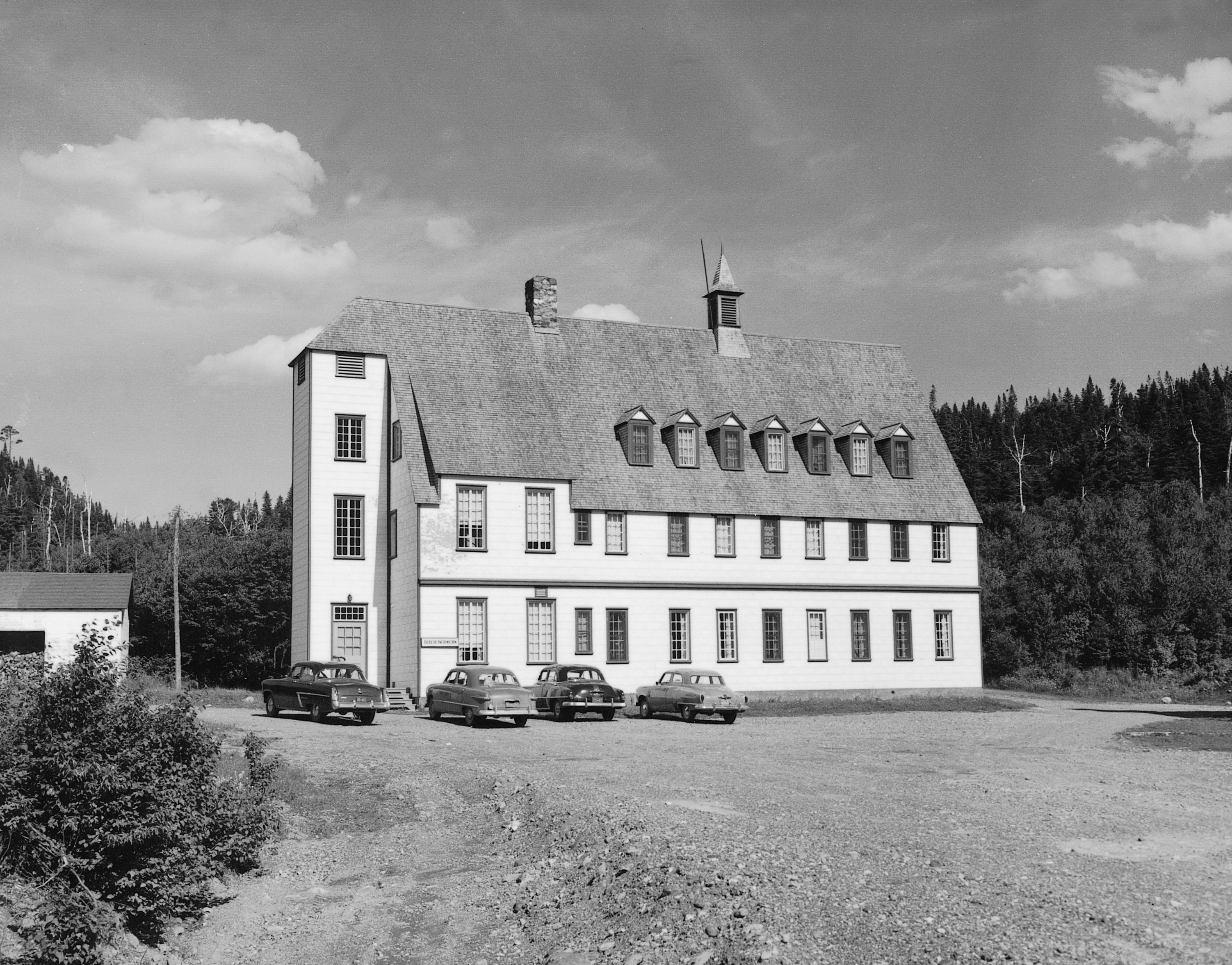 Black and white photograph of the Gîte du Mont-Albert in 1950. The inn is an imposing rectangular building of 3 stories in the heart of a coniferous forest. The pronounced angle of the double slope roof gives it an architectural style that is somewhere between that of a Swiss chalet and a manor house.