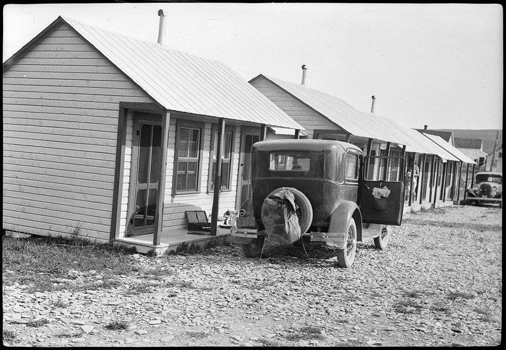 Black and white photograph of a series of tourist cabins in Percé. A 1933 car is parked in front of the first cabin.