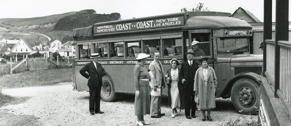 Black and white photograph of a group of people in front of a passenger bus in 1932. Behind the bus, you can see some houses in the village of Percé and the mountains that form the coast. On the bus is written Montreal Vancouver Coast to Coast New York Los Angeles.