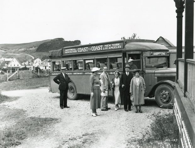 Black and white photograph of a group of people in front of a passenger bus in 1932. Behind the bus, you can see some houses in the village of Percé and the mountains that form the coast. On the bus is written Montreal Vancouver Coast to Coast New York Los Angeles.