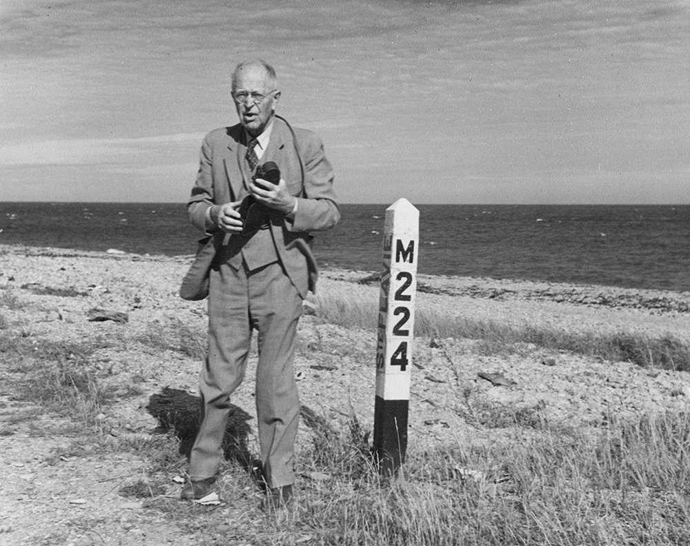 Portrait de George Parmenter. L’homme âgé, habillé en veston cravate, se tient debout, son appareil photo à la main, près d’un poteau blanc sur lequel il est inscrit M224. En arrière-plan la plage de Sainte-Flavie et la mer.