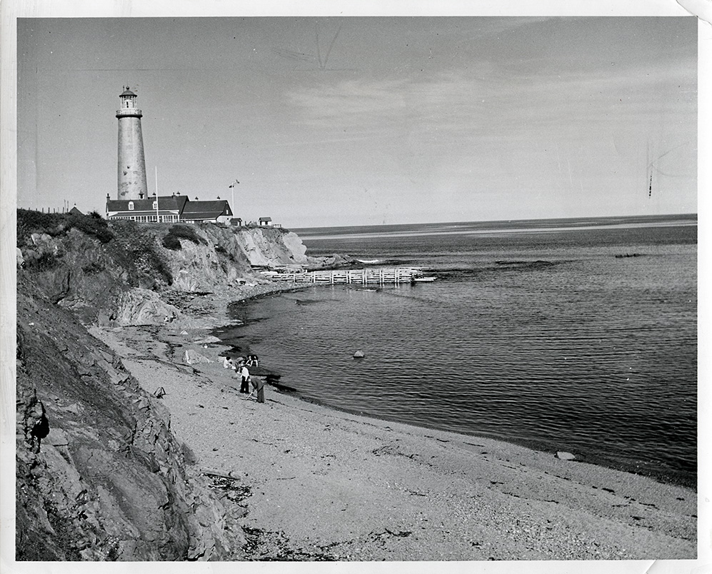 Black and white photograph of the Cap-des-Rosiers lighthouse. In a bay, a family is looking for clams on the beach. In the background, on a rocky cape, you can see the Cap-des-Rosiers lighthouse.