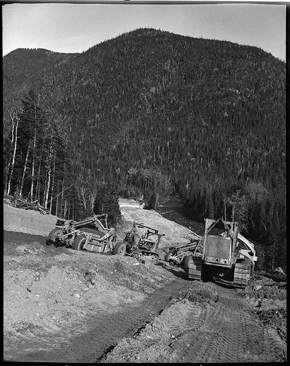 Black and white photograph of the work on the Trans-Gaspésien road. In a forest landscape, on a steep slope, construction workers operate heavy machinery to do the road preparation work.