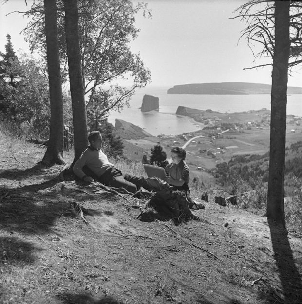 Black and white photograph of the artist Suzanne Guité working on a drawing with her husband as model, artist Alberto Tommi. The couple is sitting at the foot of a tree on top of a mountain. Behind them is a magnificent landscape at the bottom of the mountain the village of Percé as well as Percé Rock.