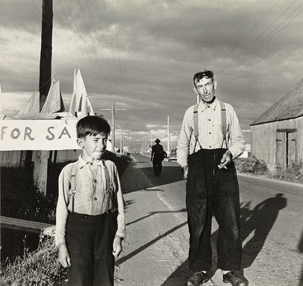 Black and white photograph of a man and a boy on the roadside. The man and the child are dressed in white shirt and black trousers held up by suspenders. Behind the boy wooden boats are placed on the top of a poster on which is written For Sale.