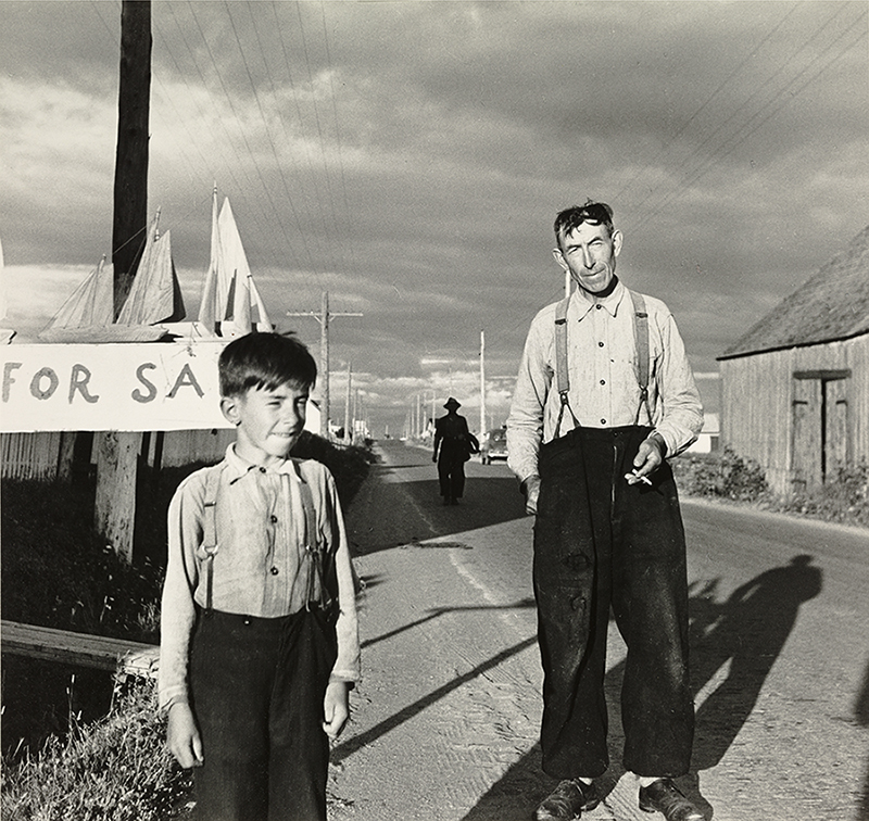 Black and white photograph of a man and a boy on the roadside. The man and the child are dressed in white shirt and black trousers held up by suspenders. Behind the boy wooden boats are placed on the top of a poster on which is written "For Sale".