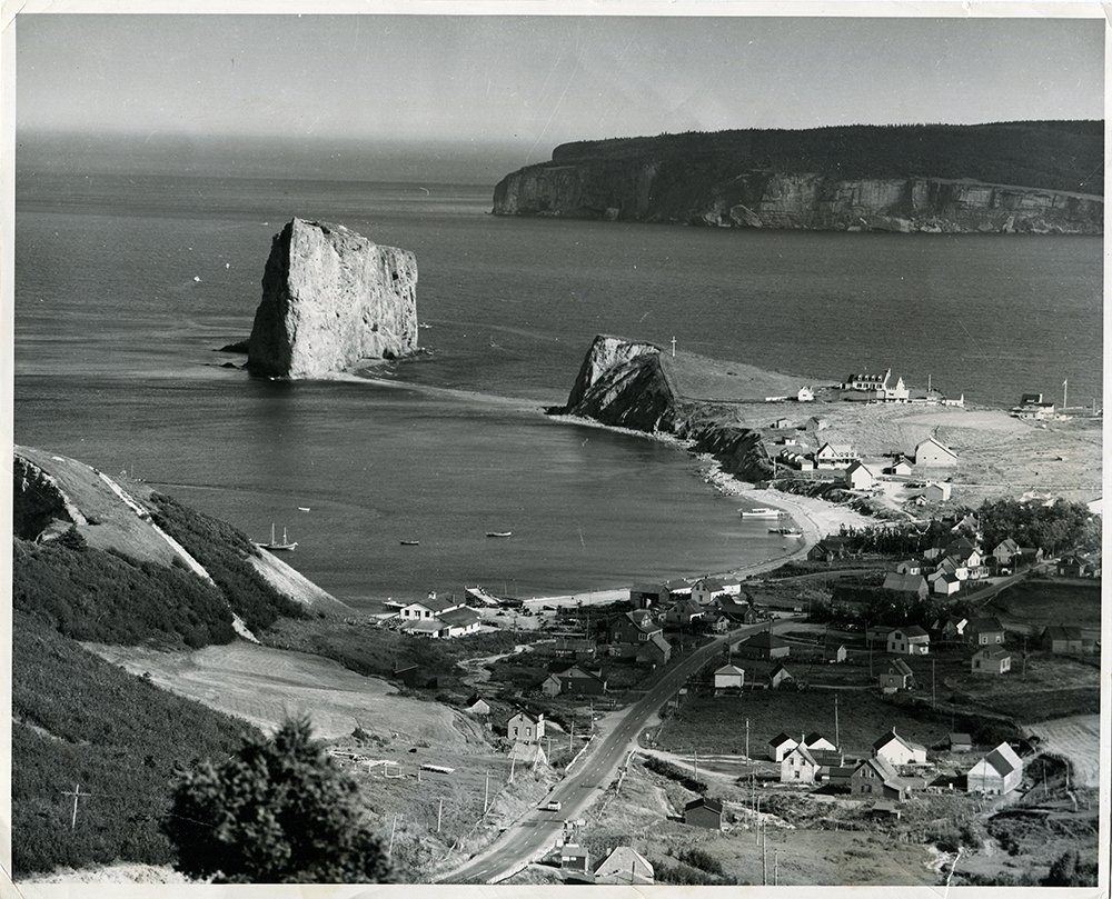 Black and white photograph from a height of land, showing the village of Percé, some fishing boats in the bay, Percé Rock and in the distance, Bonaventure Island.