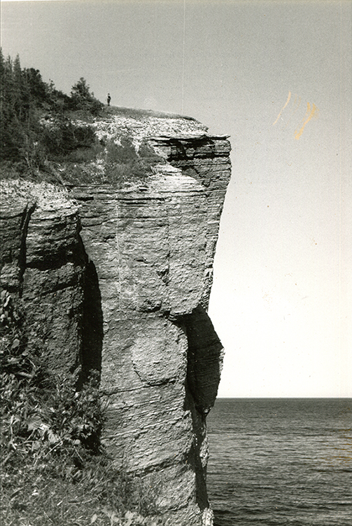 Black and white photograph of a rocky cliff of impressive height. At the top of it is the tiny silhouette of a man. In the background the vast sweep of the Gulf of St. Lawrence.