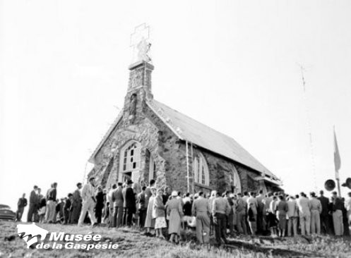 Black and white photograph of the oratory on Mont Saint-Joseph. The oratory is a stone building with a central bell tower on the front with a double slope metal roof. The oratory was built at the top of Mont Saint-Joseph. In front a crowd gathers as part of a celebration.