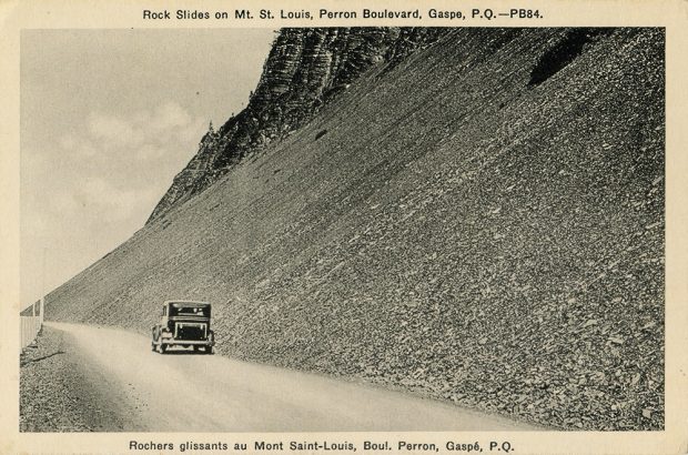 Black and white photograph of a car driving Perron Boulevard. The road follows a steep slope lined with flat rocks, leaving the impression of a recent landslide.
