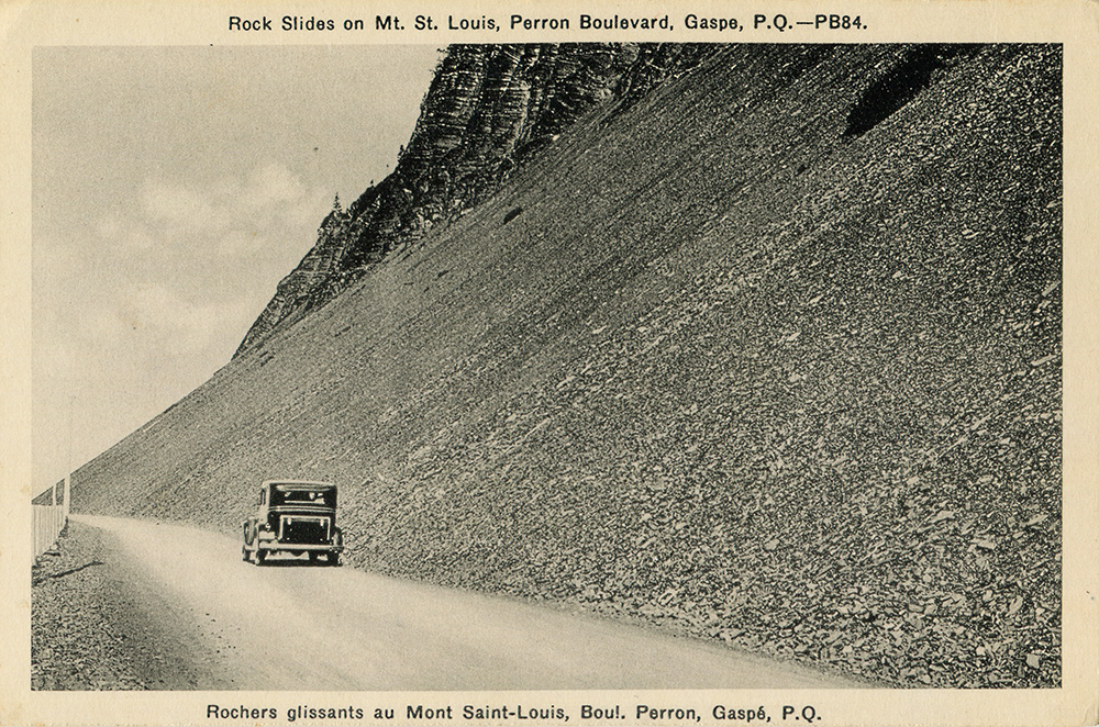 Black and white photograph of a car driving Perron Boulevard. The road follows a steep slope lined with flat rocks, leaving the impression of a recent landslide.