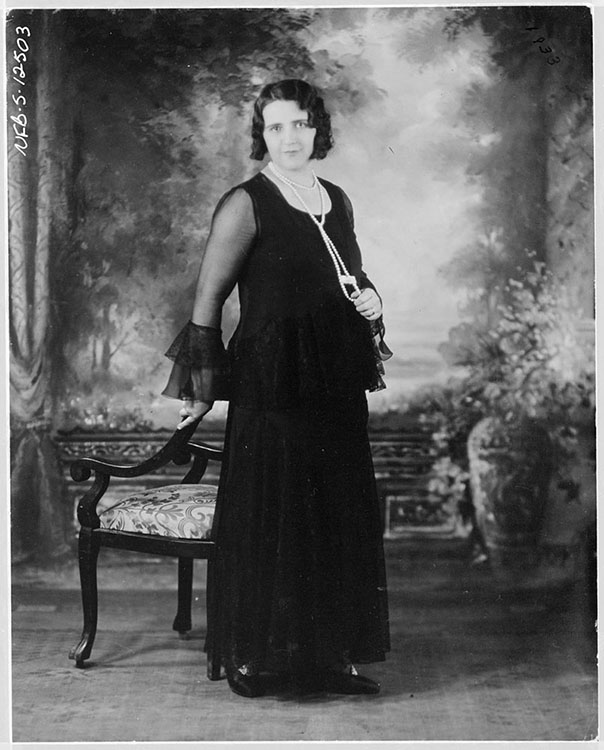 Black and white studio portrait of the singer La Bolduc. She is wearing a long black dress. She is standing in the middle of the picture, leaning on a chair in the Victorian style, in front of a painted background of classical style.