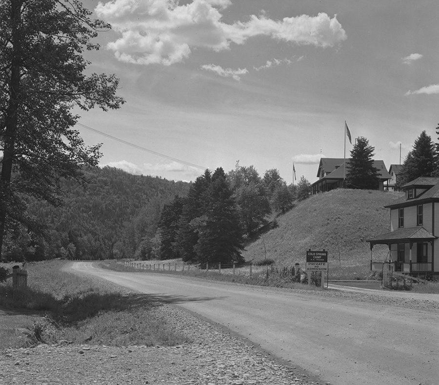 Black and white photograph taken along route 132 in the village of Routhierville in the Matapedia Valley. The road crosses a landscape of forests and mountains. On the right side of the photograph, a fishing club is perched on top of a hillock. On the side of the road sign indicates Cold Spring Camp, Private and No Admission