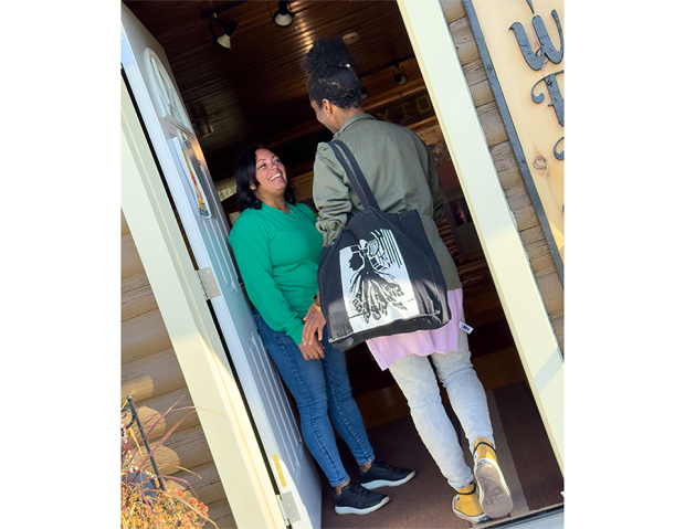 Emilie Jabouin and Michelle Robbins greeting each other in the doorway. Michelle, dressed in green, stands inside smiling; Emilie, carrying a black tote bag with a white design, stands at the entrance.