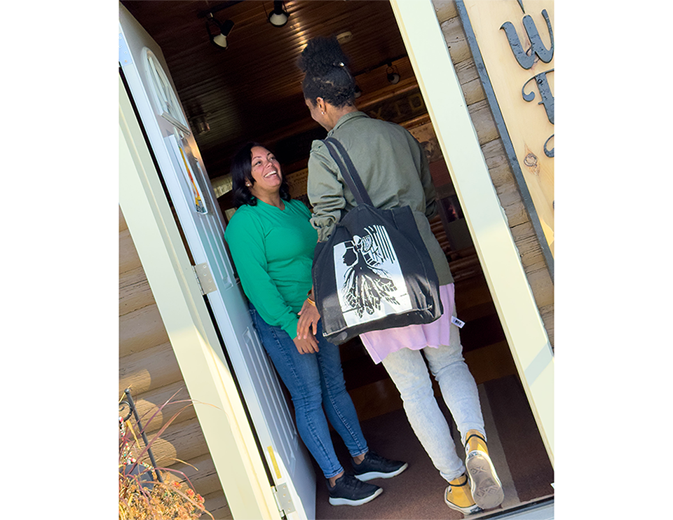 Emilie Jabouin and Michelle Robbins greeting each other in the doorway. Michelle, dressed in green, stands inside smiling; Emilie, carrying a black tote bag with a white design, stands at the entrance.
