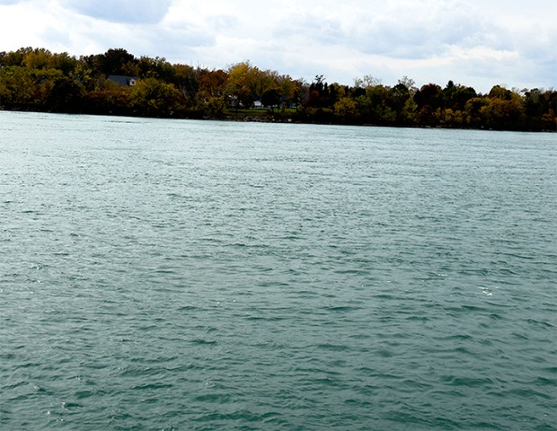 A close-up view of the Detroit River in Amherstburg, with calm water in the foreground and a shoreline lined with autumn trees in the distance under a cloudy sky. Photo by Blessing Ogunyemi.