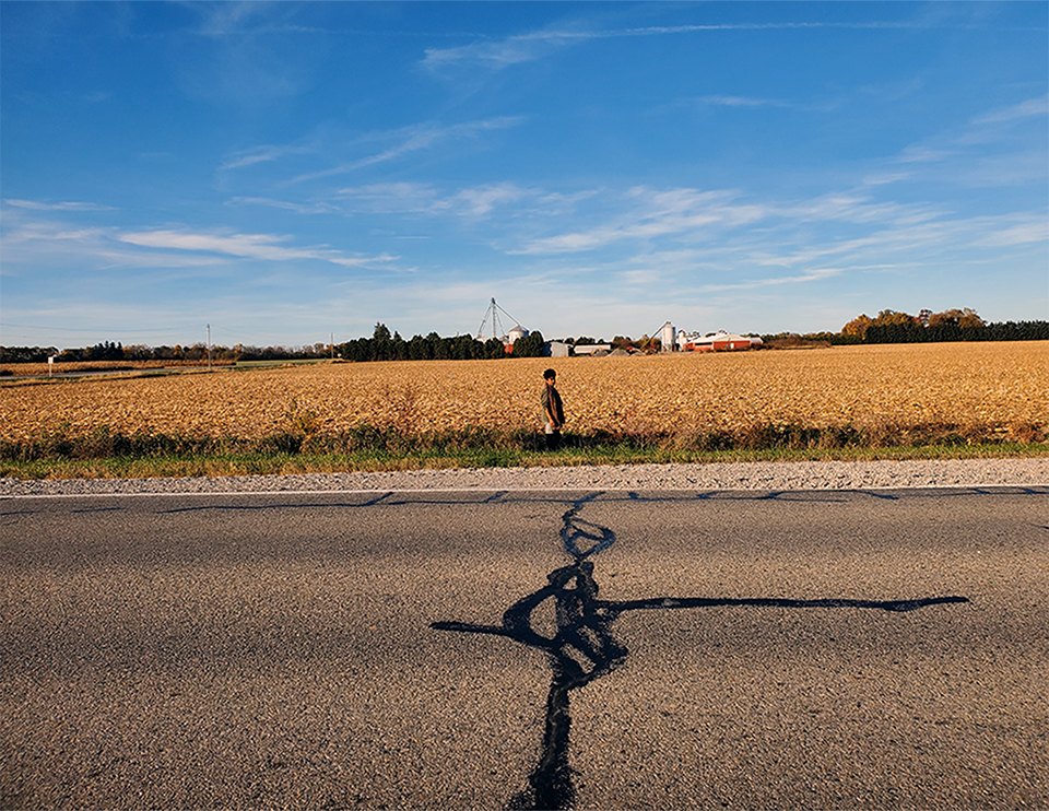 Emily standing next to a harvested field along a rural road, with farm buildings and silos visible in the background under a blue sky.