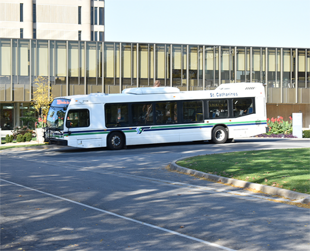 A white city bus with St. Catharines written on the side drives on a curved road in front of a building with large glass windows at Brock University