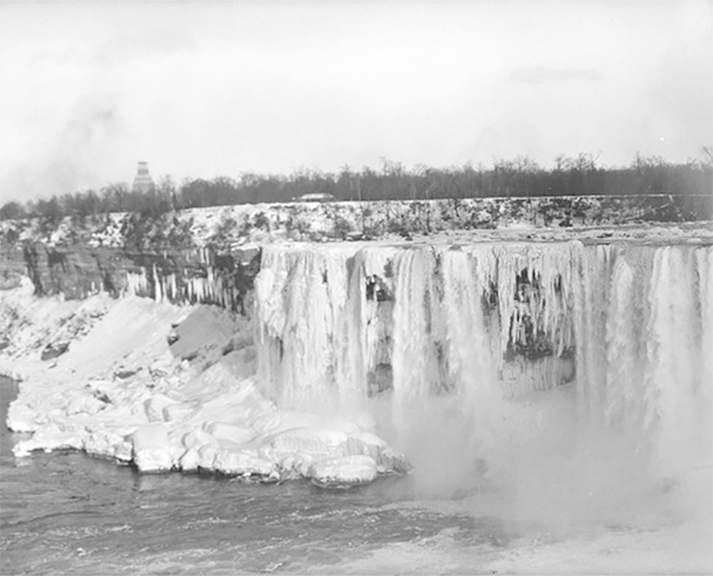 A black and white photo of Niagara Falls in winter, with large sections of the waterfall and surrounding landscape covered in ice and snow. Mist rises from the falls, and trees line the background.