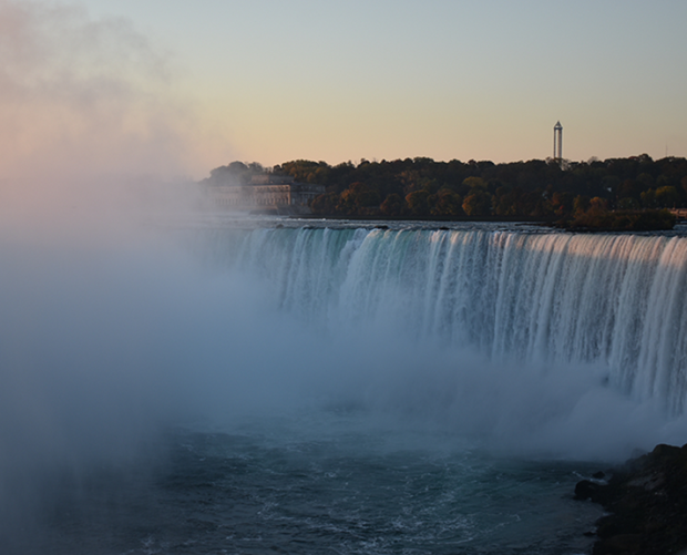 A wide view of Niagara Falls with water gushing over the edge, mist rising, and a tree-lined horizon under a clear sky at sunset.