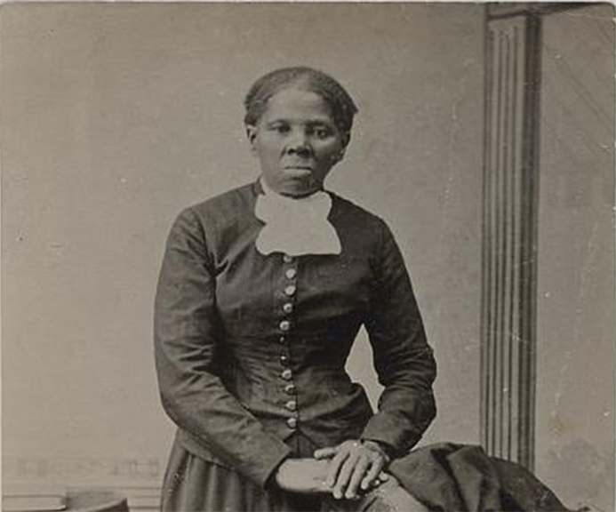 Harriet Tubman in a long, dark buttoned dress, standing beside a chair draped with a coat, with one hand resting on the chair.