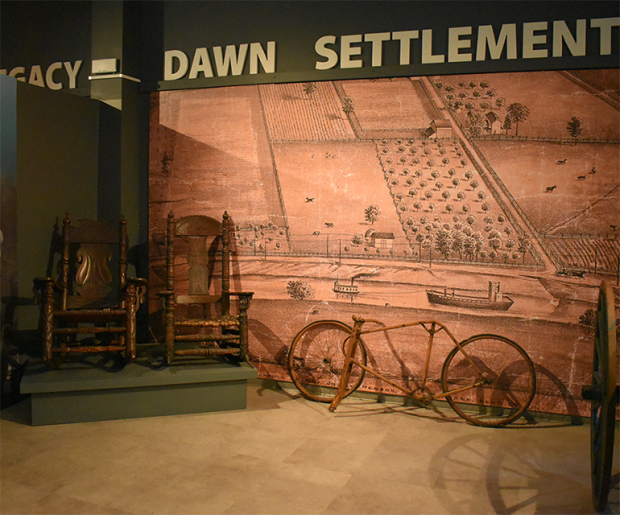 At the Josiah Henson Museum of African-Canadian History exhibit featuring two wooden rocking chairs, an old bicycle, and a mural of a rural settlement with fields and buildings. The words Dawn Settlement are displayed above the mural.