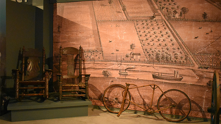 At the Josiah Henson Museum of African-Canadian History exhibit featuring two wooden rocking chairs, an old bicycle, and a mural of a rural settlement with fields and buildings. The words Dawn Settlement are displayed above the mural.