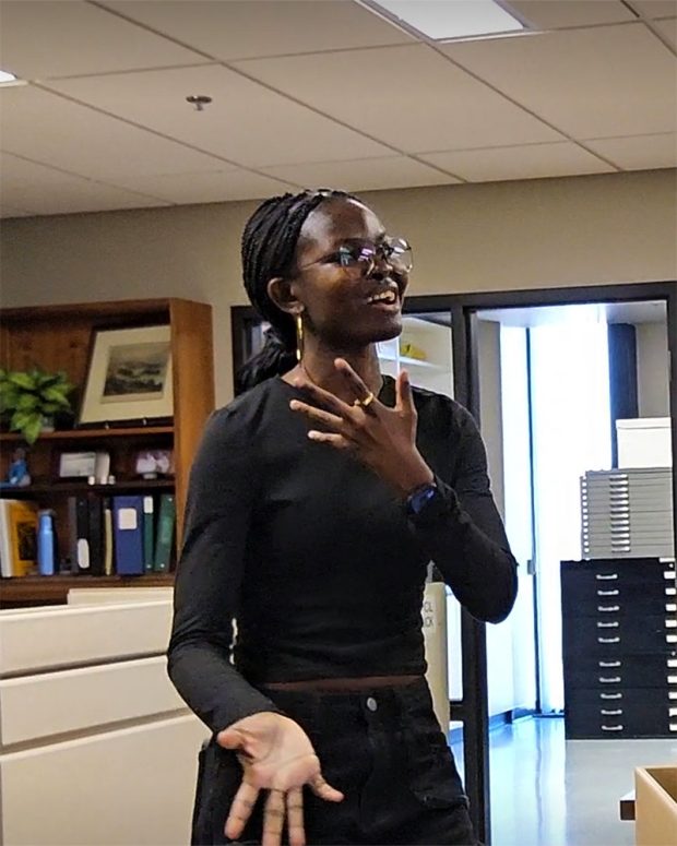 Blessings in a black long-sleeve shirt speaks expressively in an office setting. She wears glasses, earrings, and a smartwatch, with one hand raised near her chest and the other gesturing. Shelves, filing cabinets, and framed photos are visible in the background.