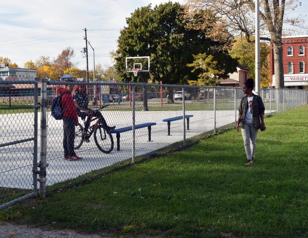 Two young men stand beside a bicycle inside a fenced basketball court on a sunny afternoon, while a young woman walks along the outside on a grassy path. In the background, autumn trees and a red brick variety store add to the neighbourhood setting.