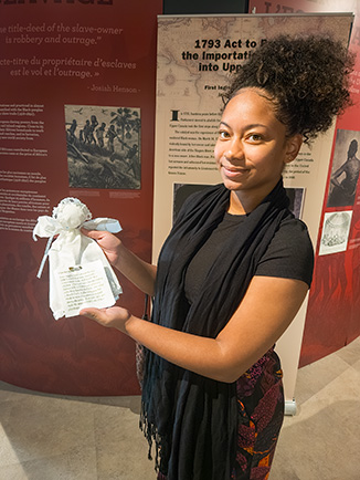 Inside the exhibition at the Josiah Henson Museum of African-Canadian History, a young woman smiles while holding a handmade handkerchief doll with a tag attached. Behind her, museum panels highlight the 1793 Act to Limit Slavery in Upper Canada and the history of slavery.