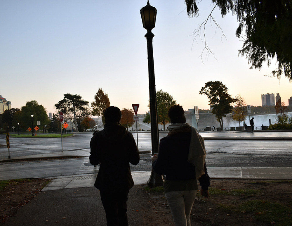 Two researchers walking along a path near a streetlamp at dawn, with trees and Niagara Falls visible in the background.