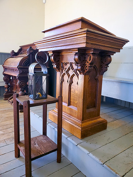 A close-up view of a carved wooden pulpit and lectern inside the historic church at the Josiah Henson Museum of African-Canadian History. A small wooden stand holds a lantern, and wooden pews line the white-painted floorboards in the background.