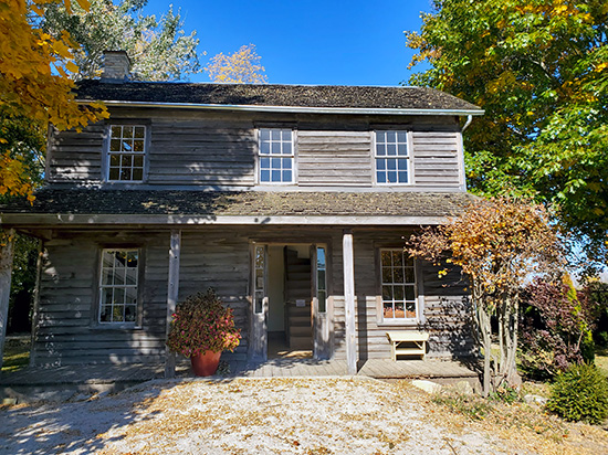 The Josiah Henson House, a two-storey log-frame home with weathered wooden siding and a covered front porch. This historic residence is part of the Josiah Henson Museum of African-Canadian History in Dresden, Ontario, surrounded by trees with early autumn foliage.