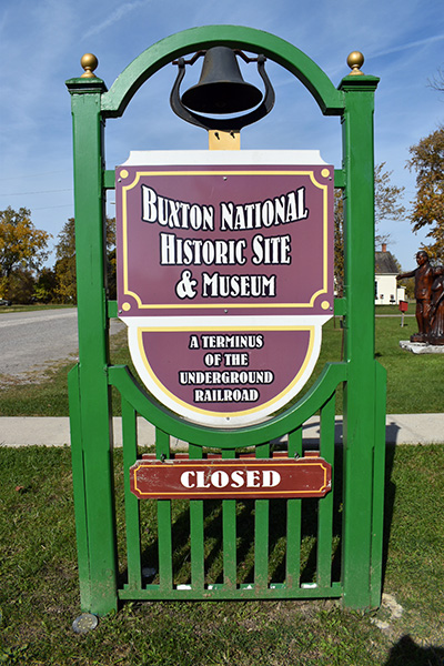 Sign for the Buxton National Historic Site & Museum, a center for preserving Black Canadian heritage. The sign is mounted on a green frame with a bell on top and a red CLOSED notice at the bottom.