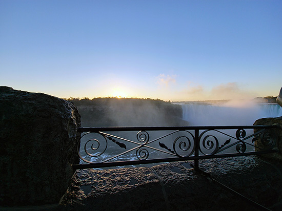 View of Niagara Falls at sunrise, with mist rising above the waterfall and sunlight glowing behind an ornate iron railing