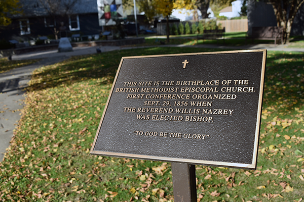 A bronze plaque mounted on a post in a park in Chatham-Kent, commemorating the birthplace of the British Methodist Episcopal Church. Fallen leaves cover the grass around it, and a historic building is visible in the background. Photo by Blessing Ogunyemi.