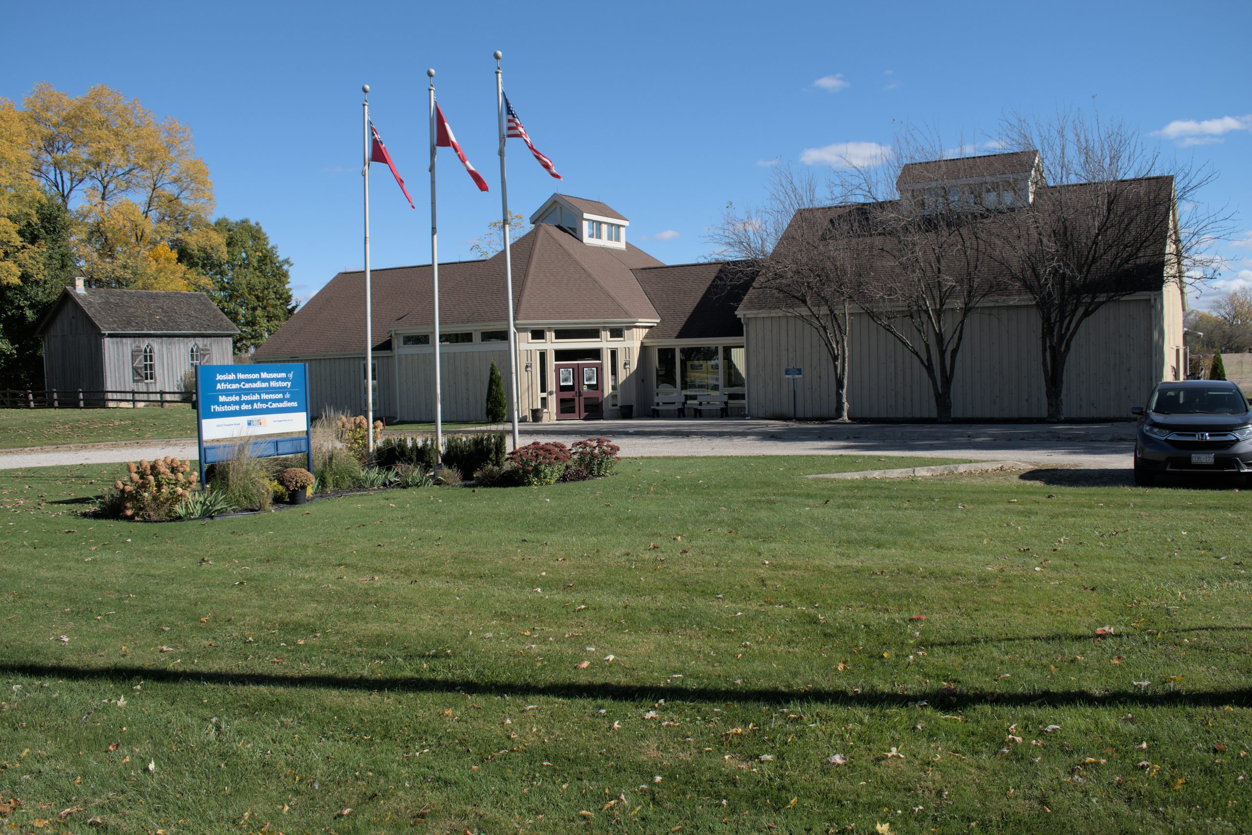 The Josiah Henson Museum of African-Canadian History on a clear day, with a row of flags flying in front, a blue museum sign, and a wide green lawn leading up to the building. Photo by Blessing Ogunyemi.