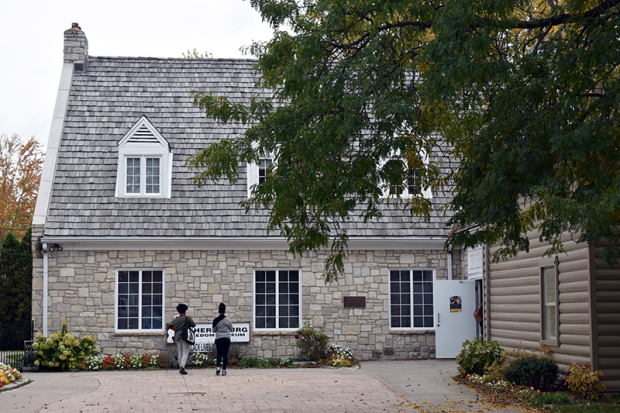 Two researchers stand outside the Amherstburg Freedom Museum, reviewing an information sign near the entrance of the historic stone building. The museum grounds are adorned with vibrant flowers and mature trees displaying autumn foliage.