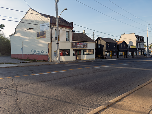 A streetscape view of Geneva Street in St. Catharines, showing a row of small storefronts and houses along the road in early morning or late-afternoon light. Power lines stretch overhead and the street appears quiet. Photo by Blessing Ogunyemi.