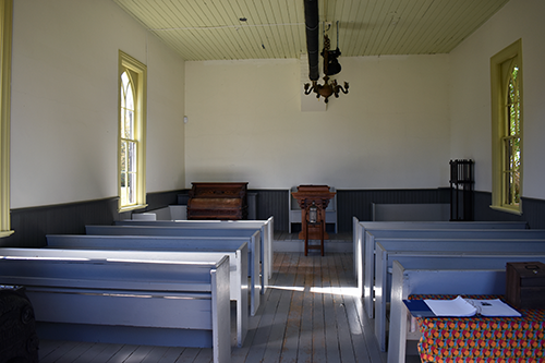 Interior of the Pioneer Church at the Josiah Henson Museum of African-Canadian History, featuring rows of simple wooden pews facing a pulpit and the original organ at the back. Sunlight streams through the tall windows on both sides.